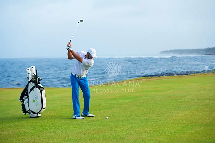 Shot of areas of the golf club with views to the ocean. A man with white shirt and blue pants swings at the ball.