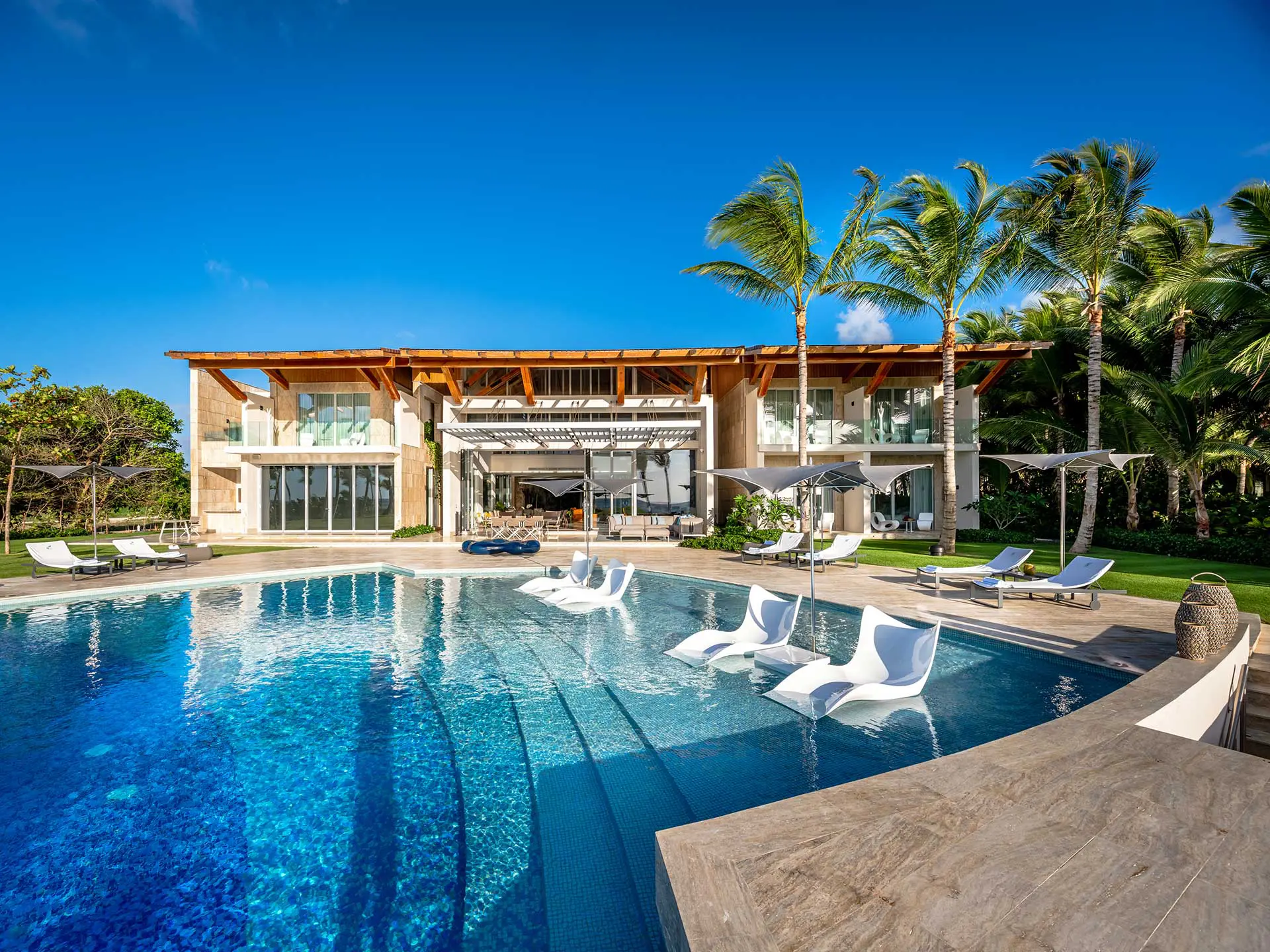 Shot looking at the front of the villa, a foliage of palm trees located in the right side blends with the structure. Visible are the balconies of five rooms that share view to the pool.